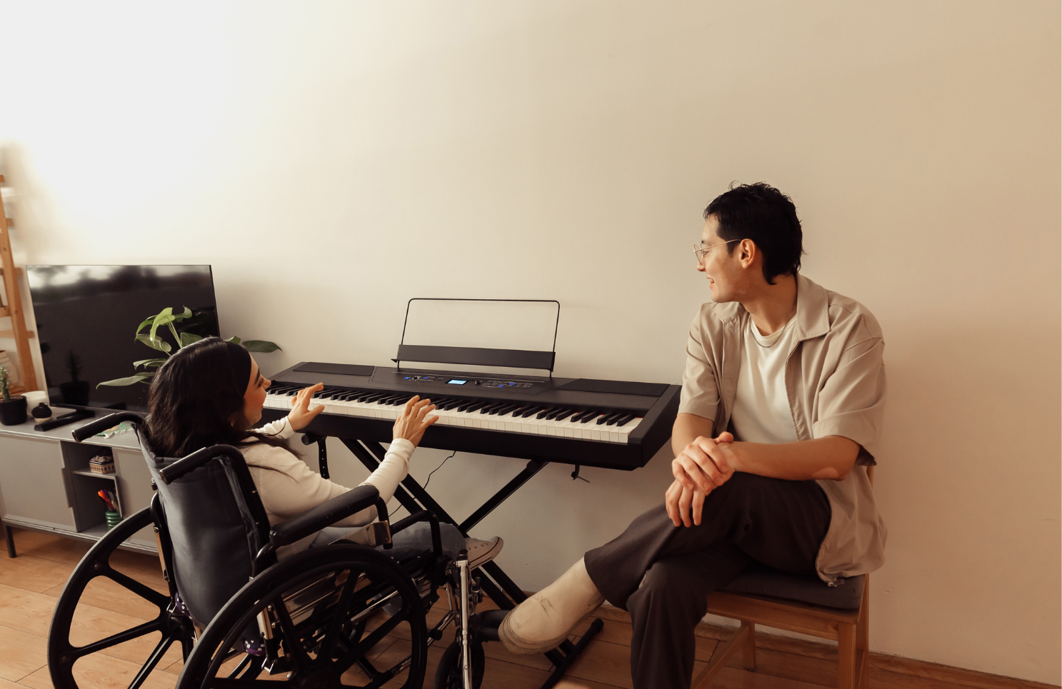 young woman in a wheelchair playing the keyboard while a man sitting on a chair chats to her